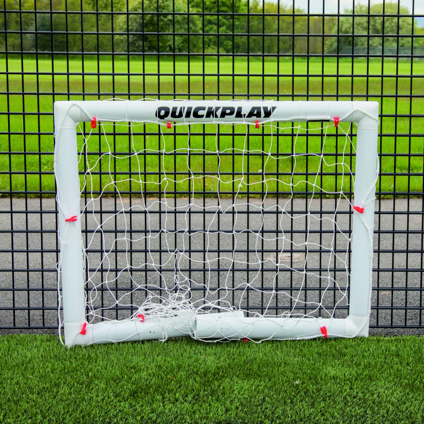 Mini-but de football Q-FOLD 90 x 75 cm plié à plat sur du gazon synthétique devant une clôture grillagée noire.