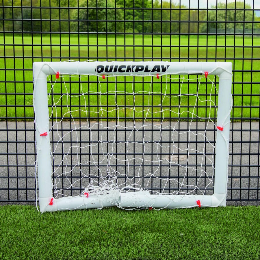 Mini-but de football Q-FOLD 90 x 75 cm plié à plat sur du gazon synthétique devant une clôture grillagée noire.