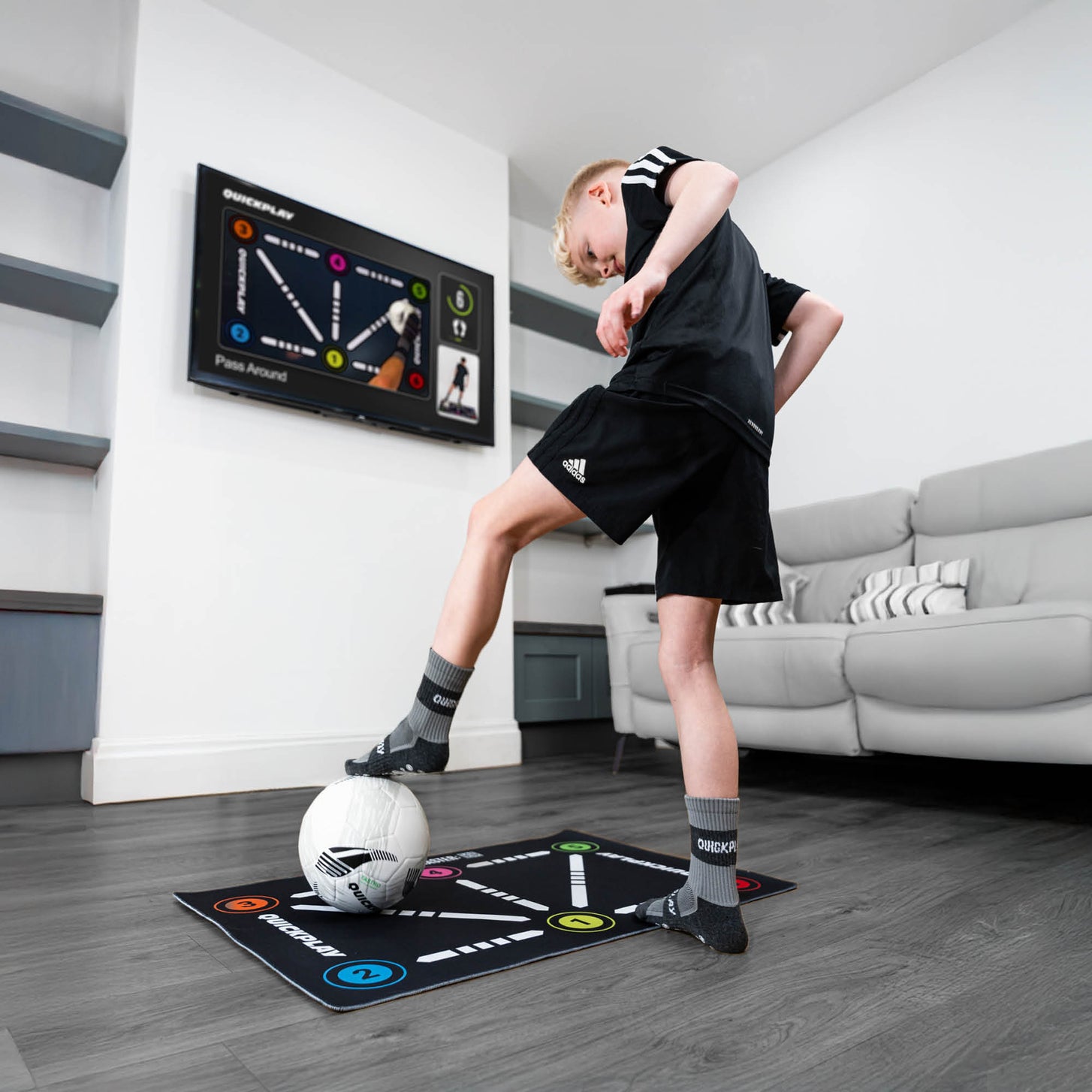 Tapis d'entraînement de football pour la maîtrise du ballon dans un salon, garçon en chaussettes grip travaillant le ballon pendant qu'une télévision diffuse un exercice.