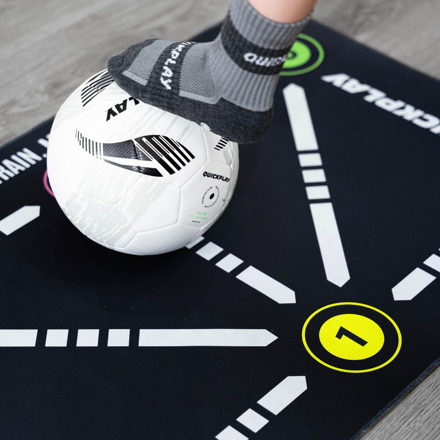 Gros plan du tapis d'entraînement de football pour la maîtrise du ballon, pied en chaussette grip QUICKPLAY sur un ballon blanc à côté du chiffre 1 jaune.