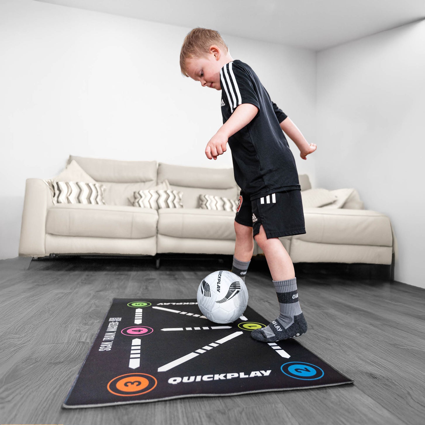 Jeune garçon travaillant le contrôle du ballon sur le tapis d'entraînement de football pour la maîtrise du ballon, sur un parquet, canapé derrière.