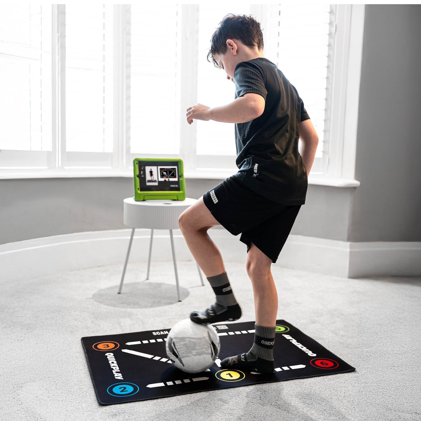 Garçon faisant un exercice sur le tapis d'entraînement de football pour la maîtrise du ballon, sur une moquette, avec une tablette affichant une vidéo près de la fenêtre.
