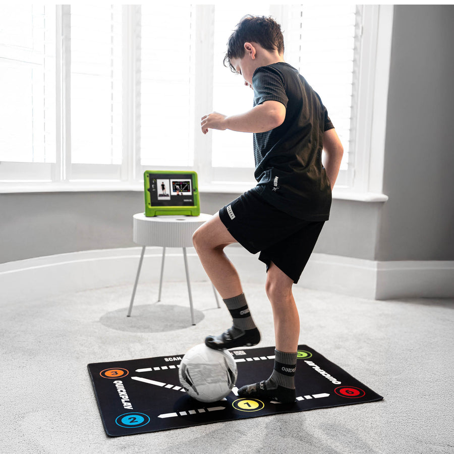 Garçon faisant un exercice sur le tapis d'entraînement de football pour la maîtrise du ballon, sur une moquette, avec une tablette affichant une vidéo près de la fenêtre.