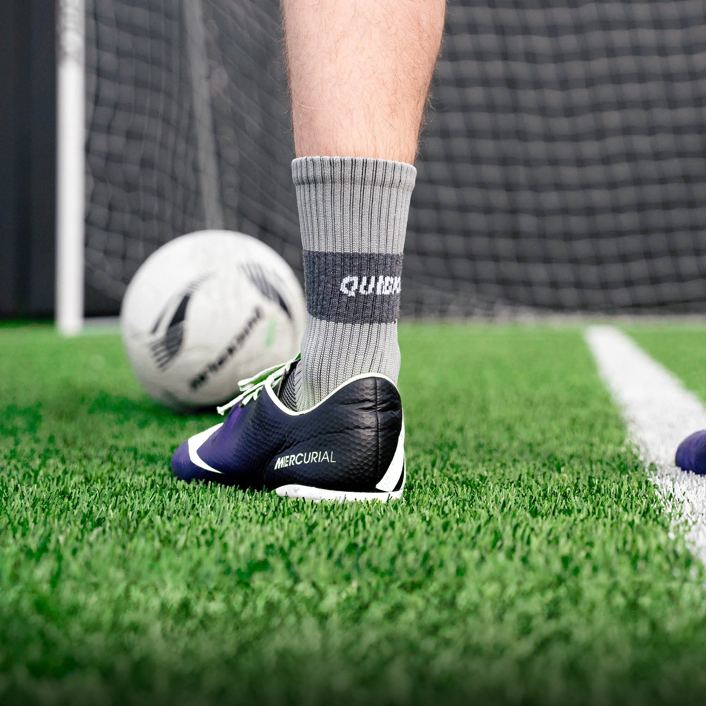 Joueur portant une chaussette grip QUICKPLAY grise avec une chaussure de football sur du gazon synthétique, ballon et filet de but derrière.