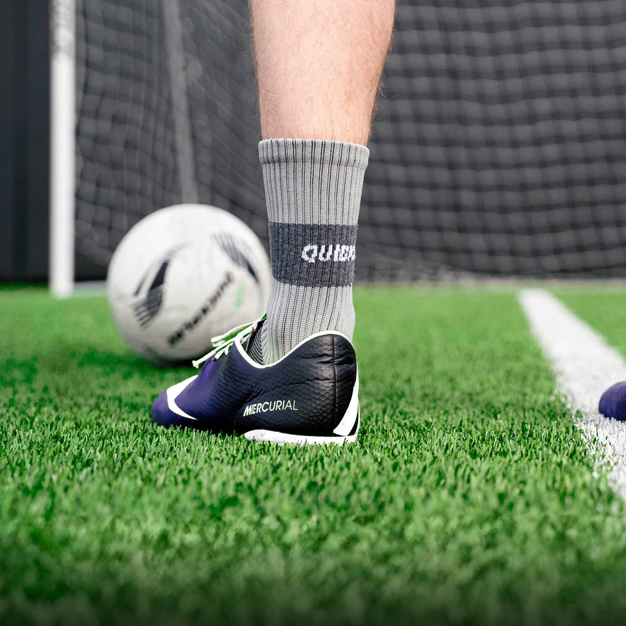 Joueur portant une chaussette grip QUICKPLAY grise avec une chaussure de football sur du gazon synthétique, ballon et filet de but derrière.