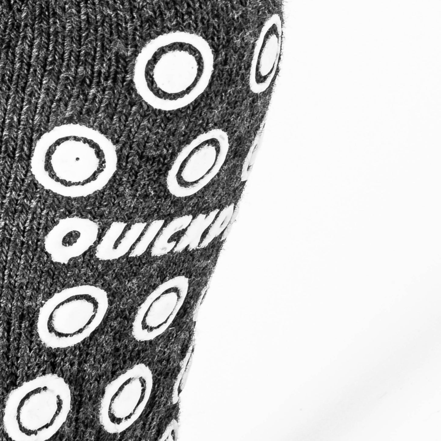Gros plan des pastilles antidérapantes blanches et du logo QUICKPLAY sur la semelle tricotée gris foncé d'une chaussette grip.
