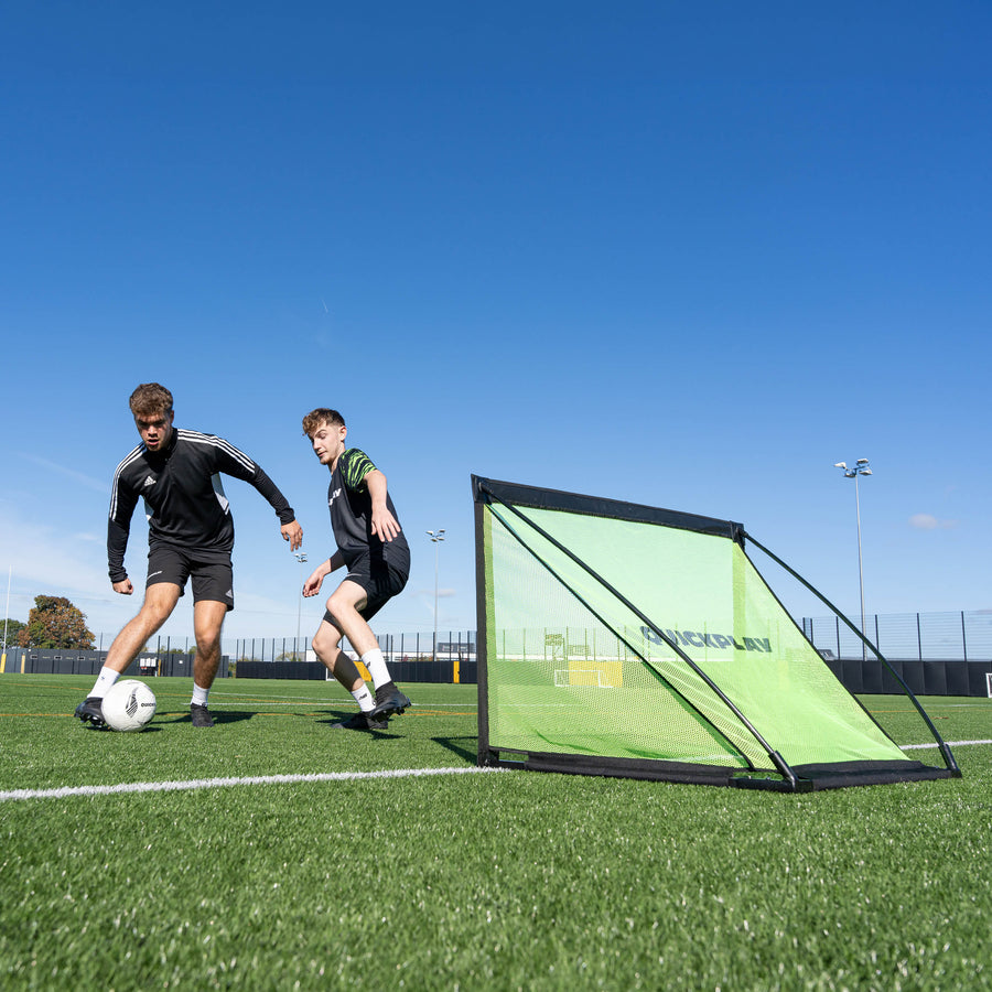 Joueurs dribblant devant le but KICKSTER PRO 120 x 75 cm sur un terrain synthétique par une journée ensoleillée.