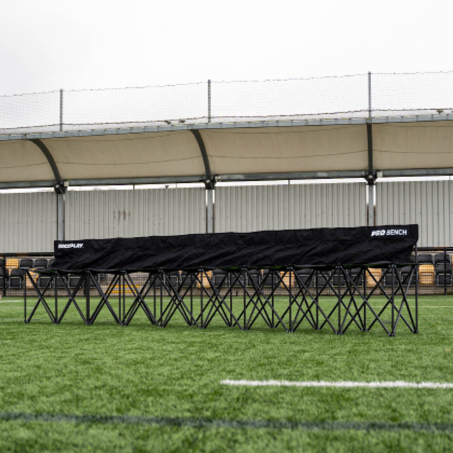 Long banc de touche PRO Bench 9 places noir, déplié sur du gazon synthétique devant une tribune couverte.