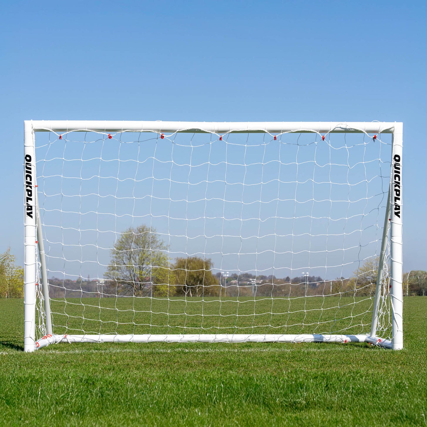 But de football Q-FOLD 240 x 150 cm vu de face sur un terrain en herbe, avec des arbres visibles à travers le filet blanc.