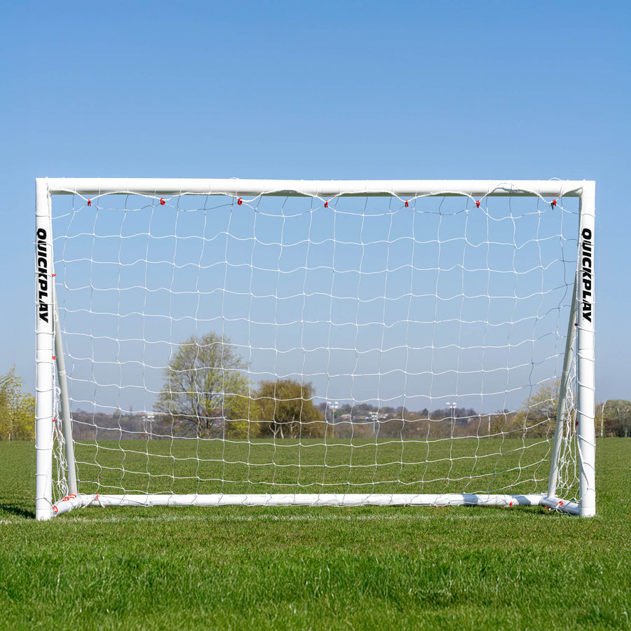 But de football Q-FOLD 240 x 150 cm vu de face sur un terrain en herbe, avec des arbres visibles à travers le filet blanc.