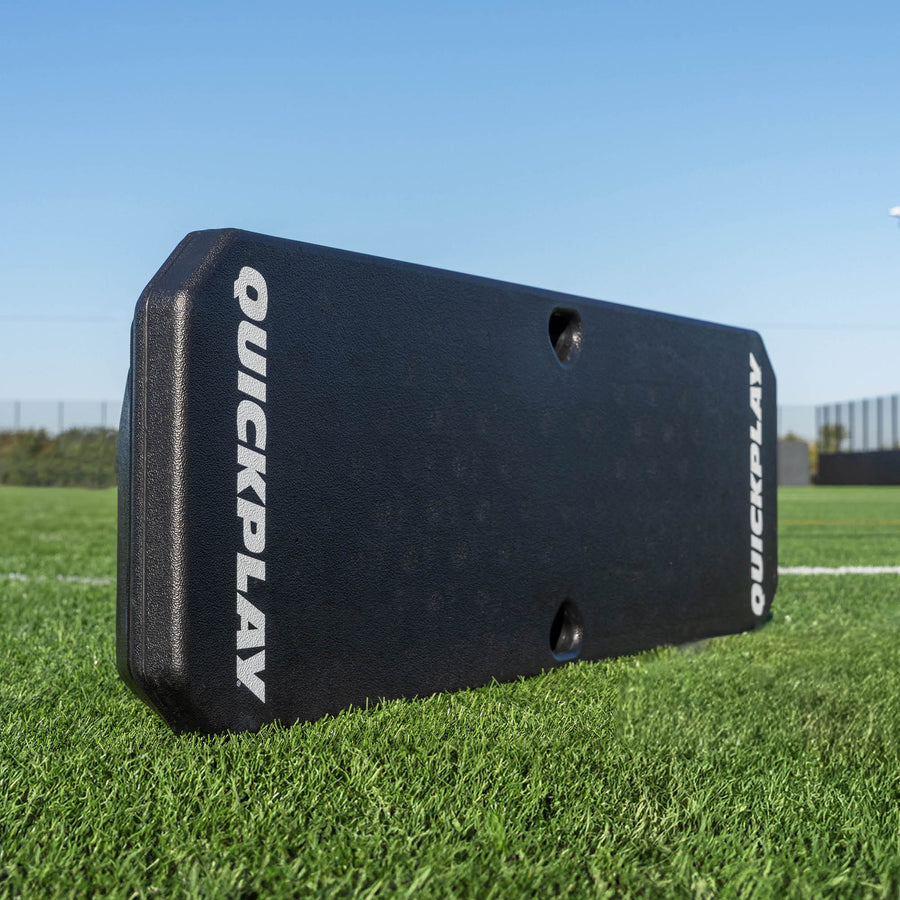 Planche de rebond de football REPLAY Boombox, noire avec des logos QUICKPLAY blancs, posée sur du gazon synthétique.