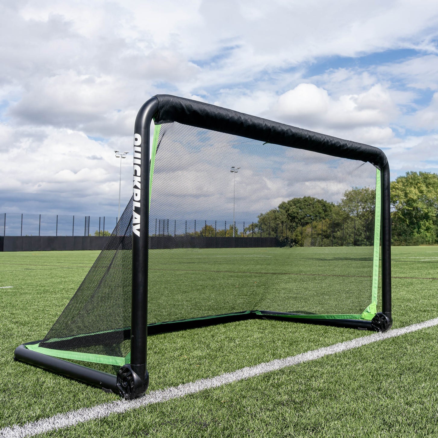 But de football RHINO 150 x 100 cm installé sur un terrain synthétique sous un ciel nuageux, avec des arbres derrière la clôture.
