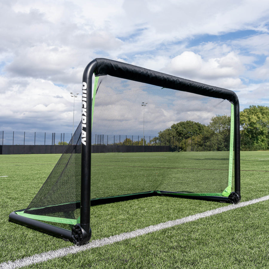 But de football RHINO 150 x 100 cm installé sur un terrain synthétique sous un ciel nuageux, avec des arbres derrière la clôture.