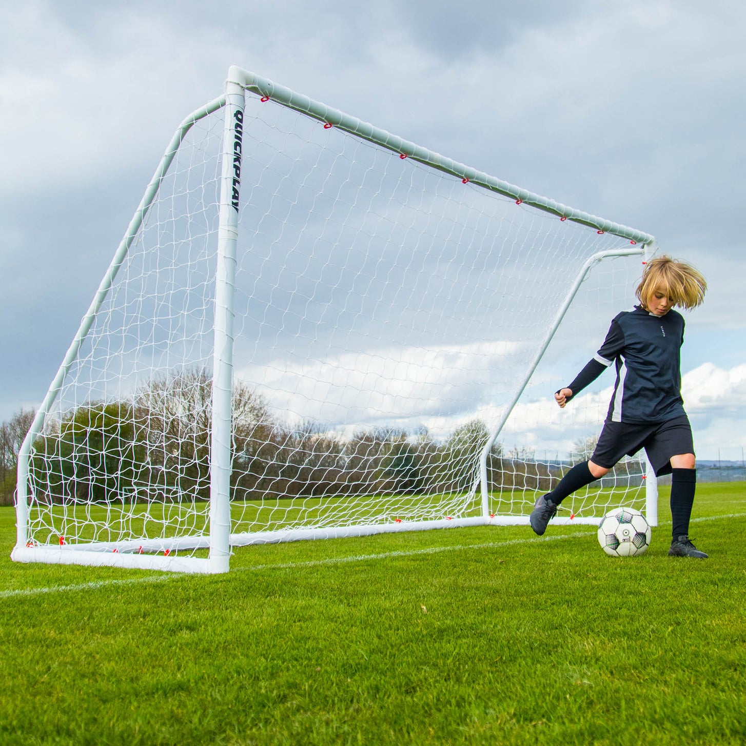 Enfant dribblant devant un but Q-FOLD Match blanc sur un terrain en herbe sous un ciel nuageux.