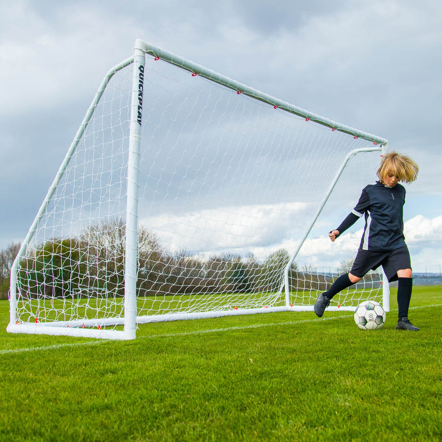 Enfant dribblant devant un but Q-FOLD Match blanc sur un terrain en herbe sous un ciel nuageux.