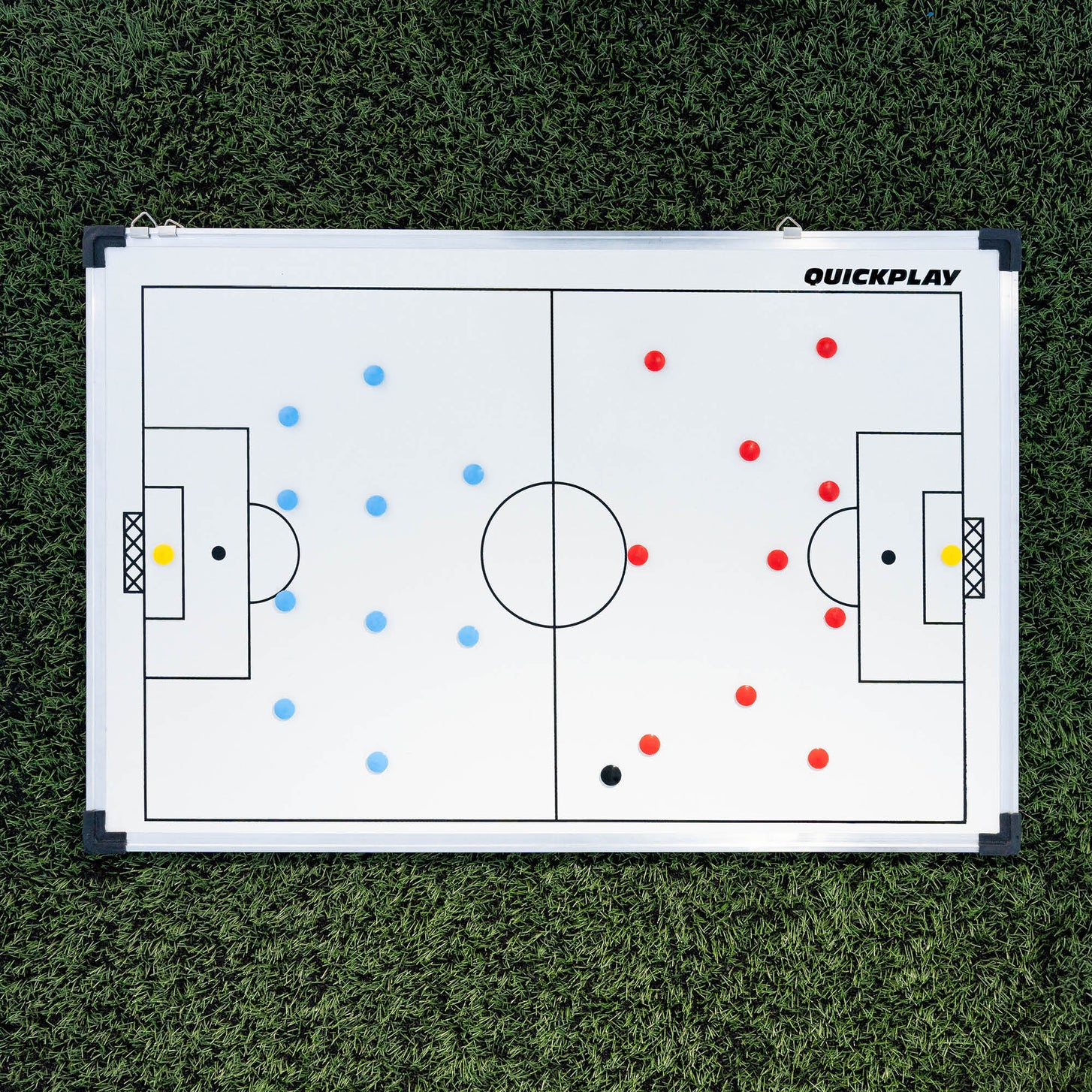 Tableau tactique de football magnétique 60 x 90 cm posé à plat sur du gazon synthétique, avec deux équipes d'aimants rouges et bleus en place.