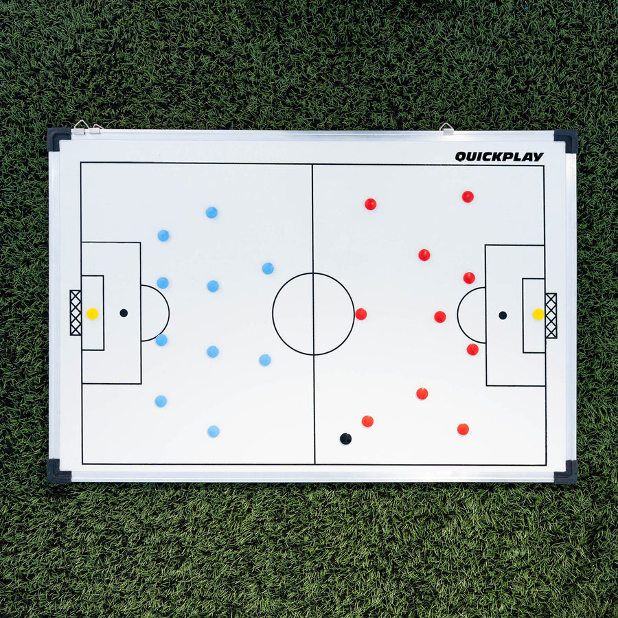 Tableau tactique de football magnétique 60 x 90 cm posé à plat sur du gazon synthétique, avec deux équipes d'aimants rouges et bleus en place.