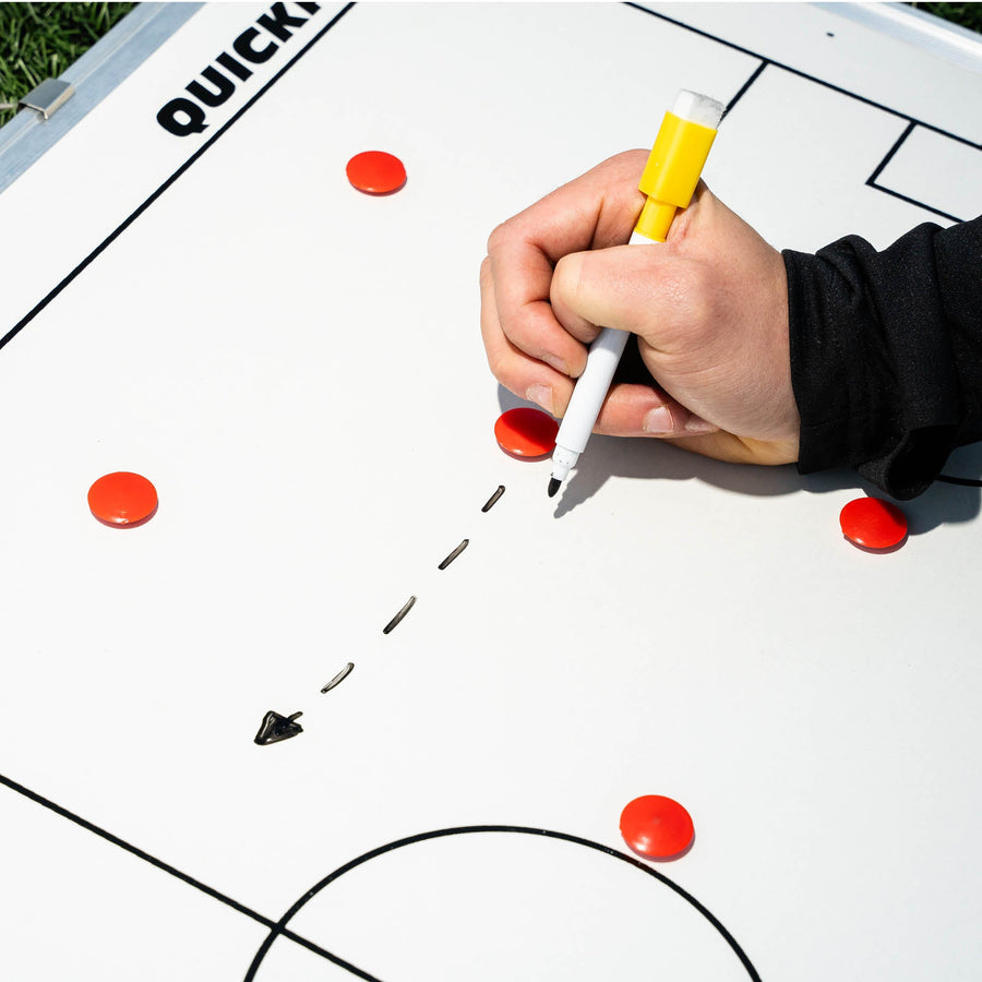 Tableau tactique de football magnétique 60 x 90 cm en cours d'utilisation : une main trace une flèche en pointillés au feutre jaune parmi des aimants rouges.