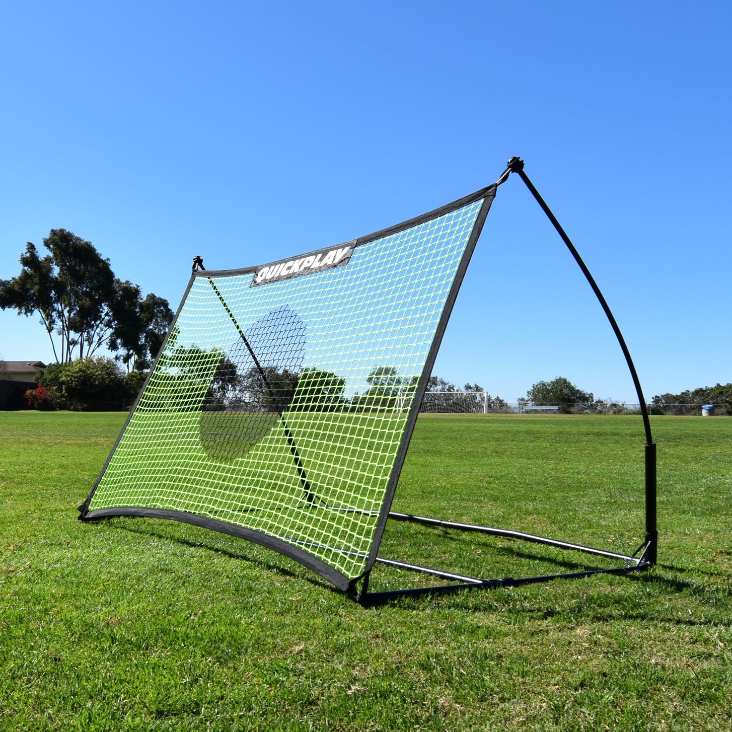 Rebondisseur de football TEKKERS Net 150 x 100 cm sur l'herbe au soleil, vu depuis un angle avant, avec son filet vert.