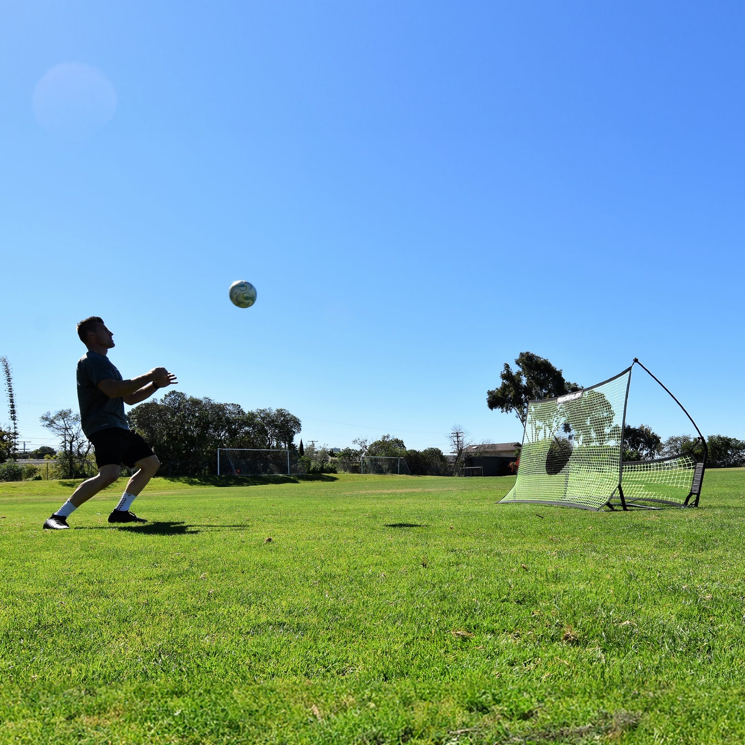 Vue large d'un joueur se préparant à contrôler un ballon en l'air, avec le rebondisseur TEKKERS Trainer derrière lui sur l'herbe.