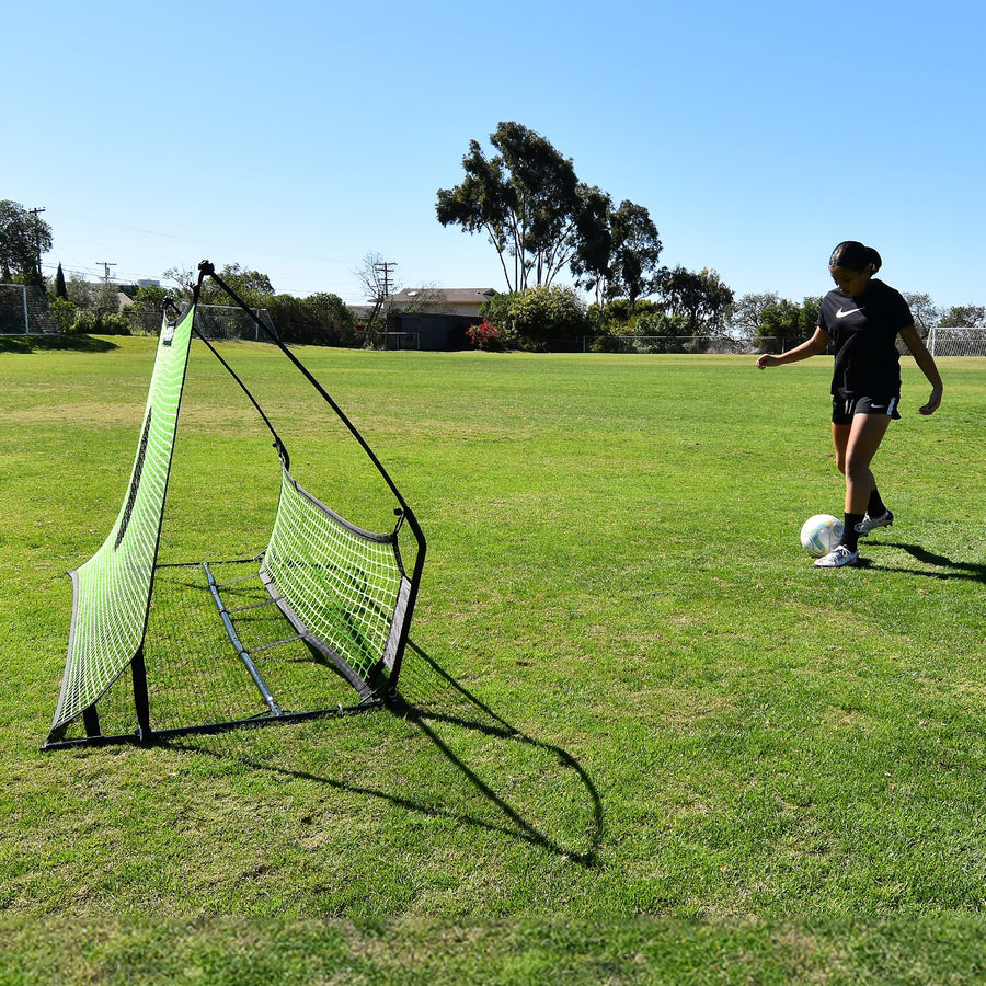 Joueur sur le point de passer un ballon dans le rebondisseur de football à double filet TEKKERS Trainer, vu de côté sur l'herbe.